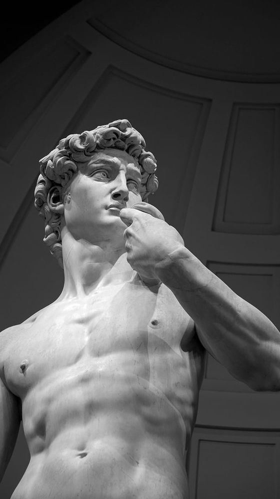 Marble statue of a muscular male figure with curly hair, gazing thoughtfully, set against a curved architectural background.