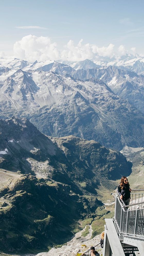 A person stands on a viewing platform overlooking a vast mountainous landscape with rugged peaks and valleys under a cloudy sky.