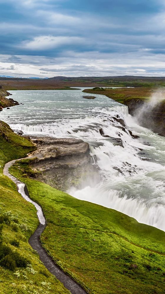 Aerial view of Gullfoss waterfall, cascading over rocky cliffs with lush green surroundings and a cloudy sky in Iceland.