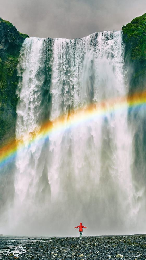 Person in a red jacket standing in front of a large waterfall with a rainbow arching across the misty water.