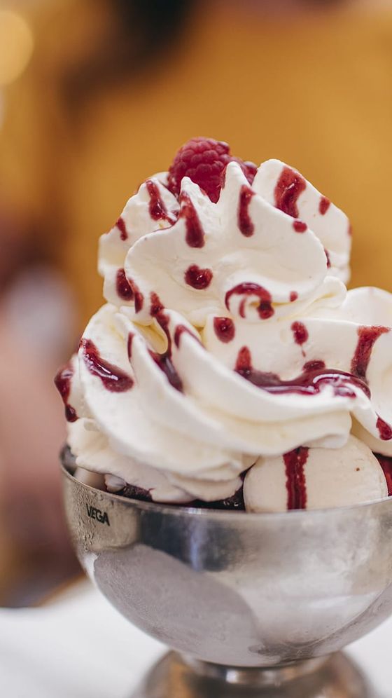 A metal bowl filled with swirled vanilla ice cream topped with whipped cream, raspberry sauce, and a single raspberry.