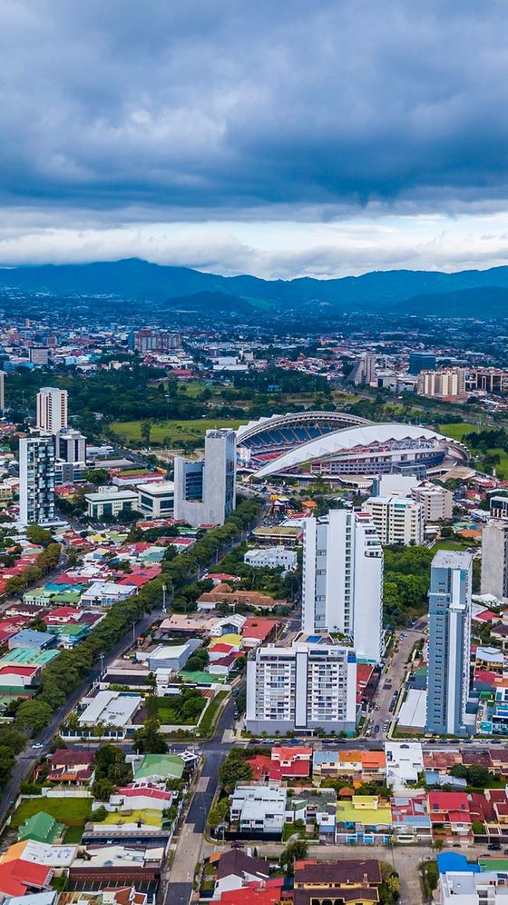 Aerial view of a cityscape featuring residential buildings, a stadium, green spaces, and mountains under a cloudy sky.