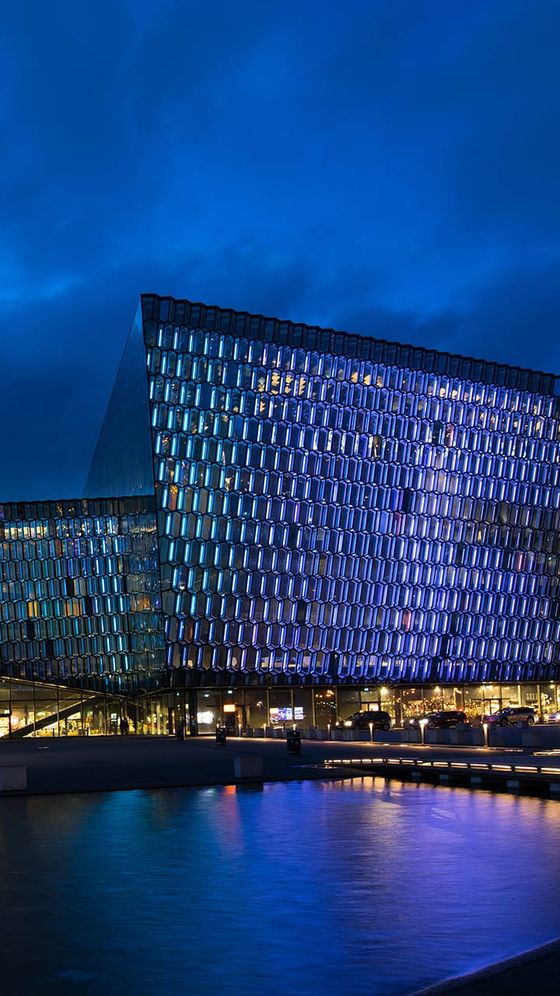Modern glass building with illuminated facade at dusk, reflecting vibrant blue and purple hues on a nearby water surface under a cloudy sky.
