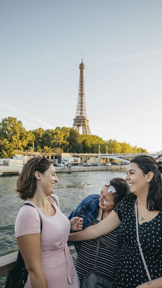 Three women laughing together by the Seine River with the Eiffel Tower in the background on a clear day.