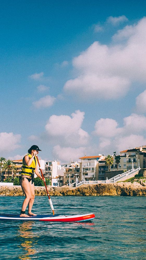 Person paddleboarding on calm water near rocky shore, under a blue sky with fluffy clouds, and buildings in the background.