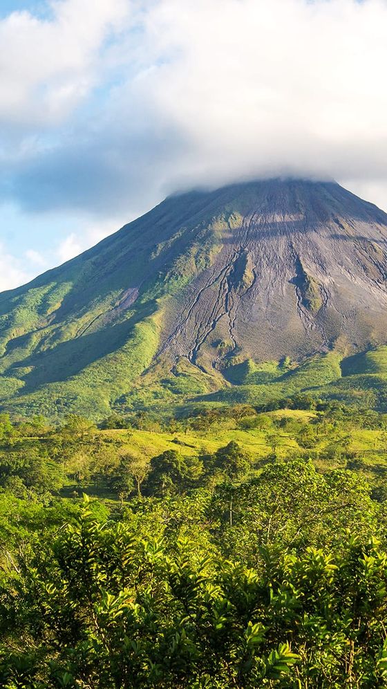 A scenic view of a lush green landscape with a towering volcano under a partly cloudy sky.
