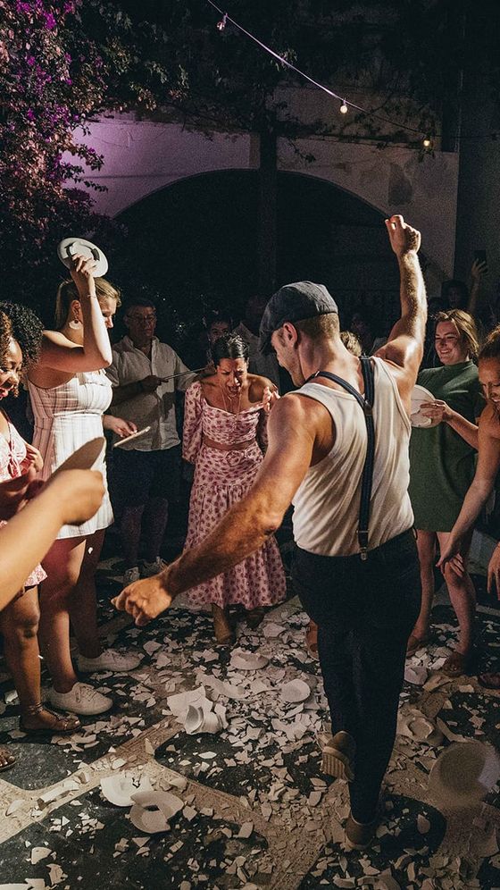 A lively party scene with people dancing; broken plates scattered on the ground, and string lights above create a festive atmosphere.