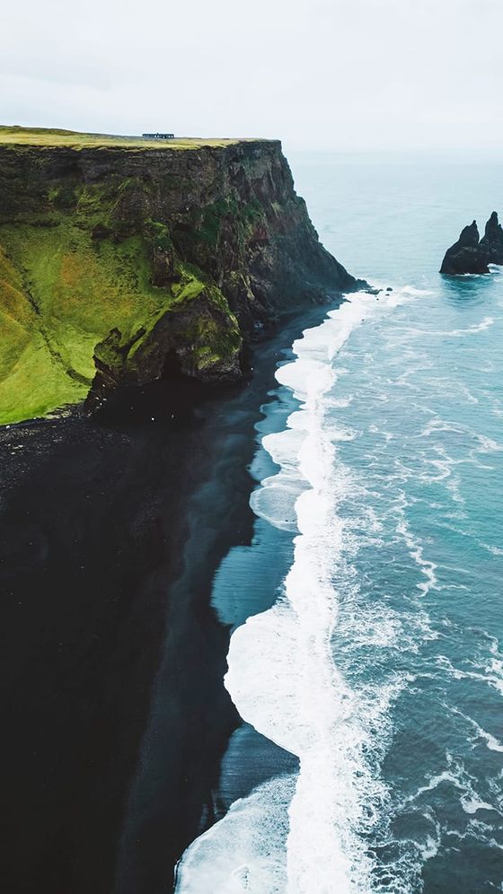 Aerial view of a black sand beach with crashing waves, steep green cliffs, and a distant house under a cloudy sky.