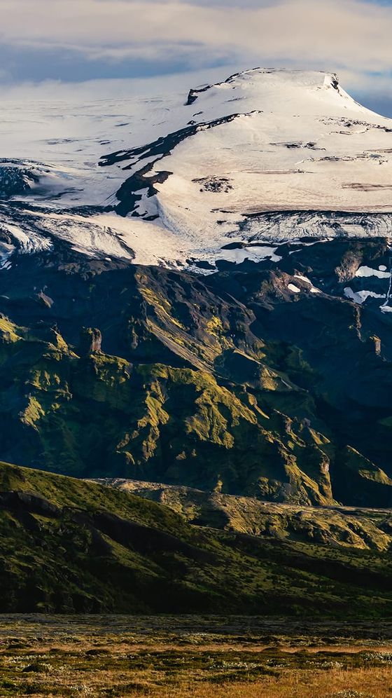 Snow-capped mountain with rocky ridges, lush green vegetation, and a cloudy sky.