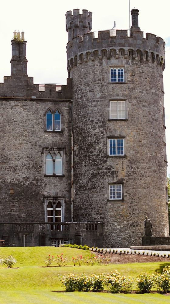 Stone castle with a cylindrical tower featuring decorative battlements, small windows, and a manicured garden with blooming roses in front.