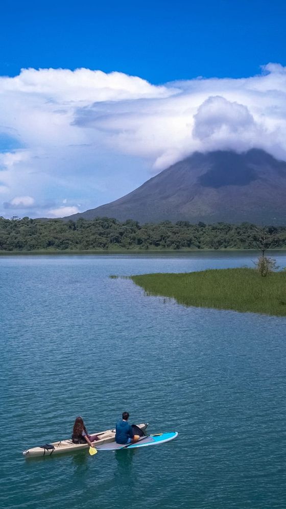 Two people kayaking on a tranquil lake with a lush green shoreline and a mist-covered volcano in the background under a blue sky.