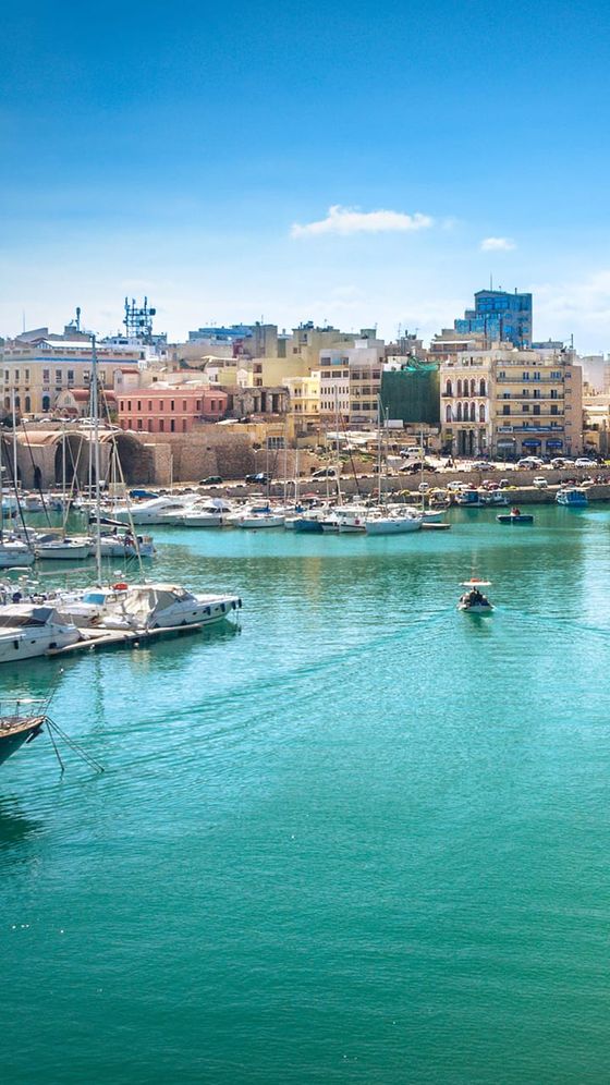 Scenic harbor view with boats on calm teal water, surrounded by colorful buildings under a clear blue sky.