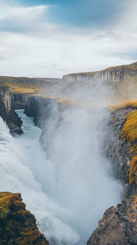 A powerful waterfall cascades into a misty gorge surrounded by rugged cliffs and grassy terrain under a cloudy blue sky.