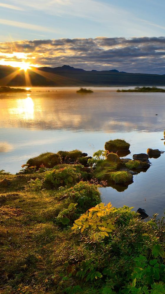 Sunset over a serene lake with patches of green grass and small rocks in the foreground, silhouetted mountains, and a cloudy sky.