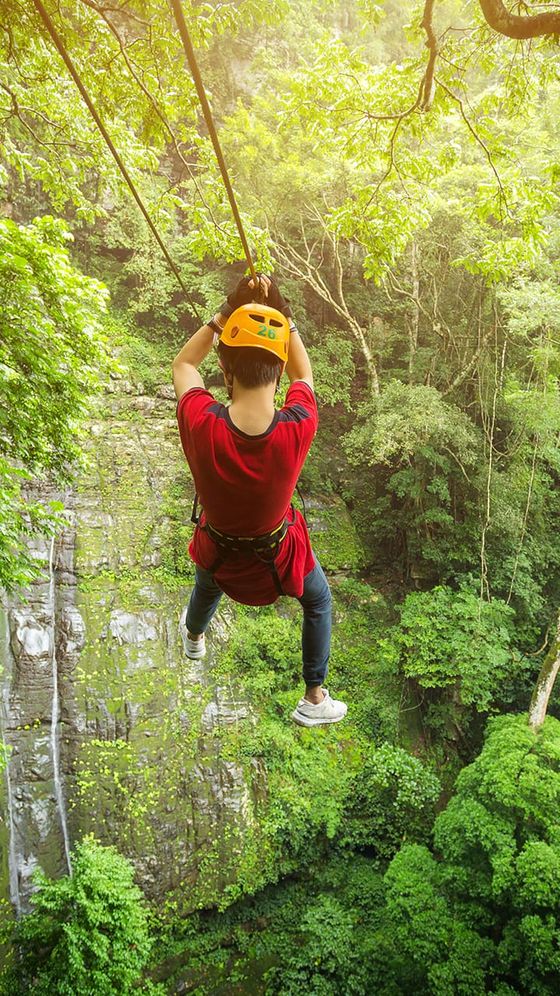 A person in a red shirt and helmet ziplining over a lush green forest with a cliff in the background.