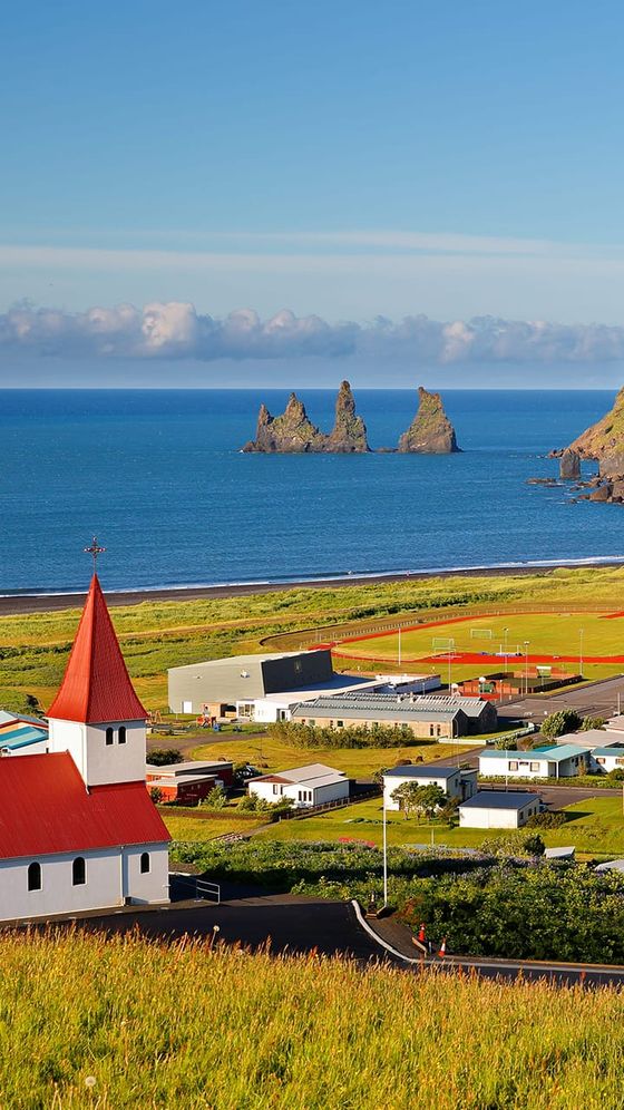 A coastal village with a red-roofed church, fields, and rocky sea stacks under a clear blue sky.