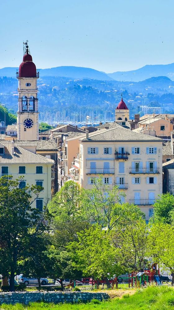 Scenic view of Corfu town with a prominent clock tower, surrounded by traditional buildings, lush trees, and mountains in the background.