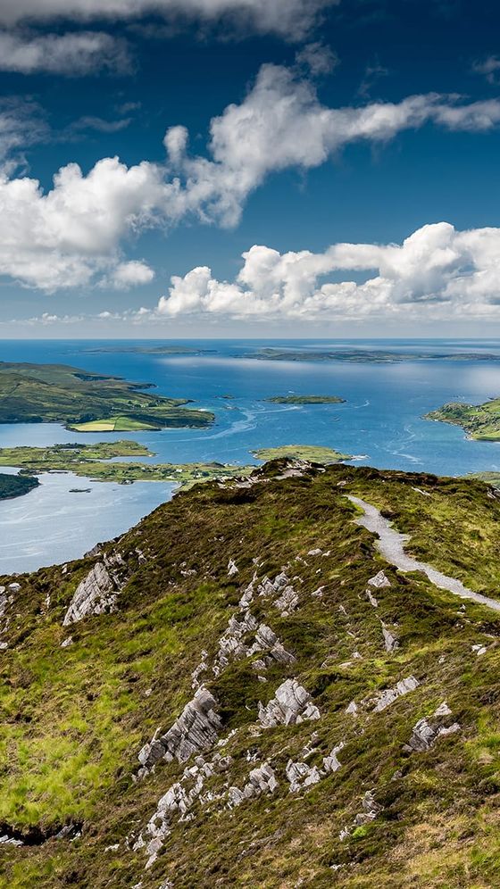 Scenic view from a rocky hilltop overlooking a lush landscape with a winding path, lakes, and a cloudy blue sky.