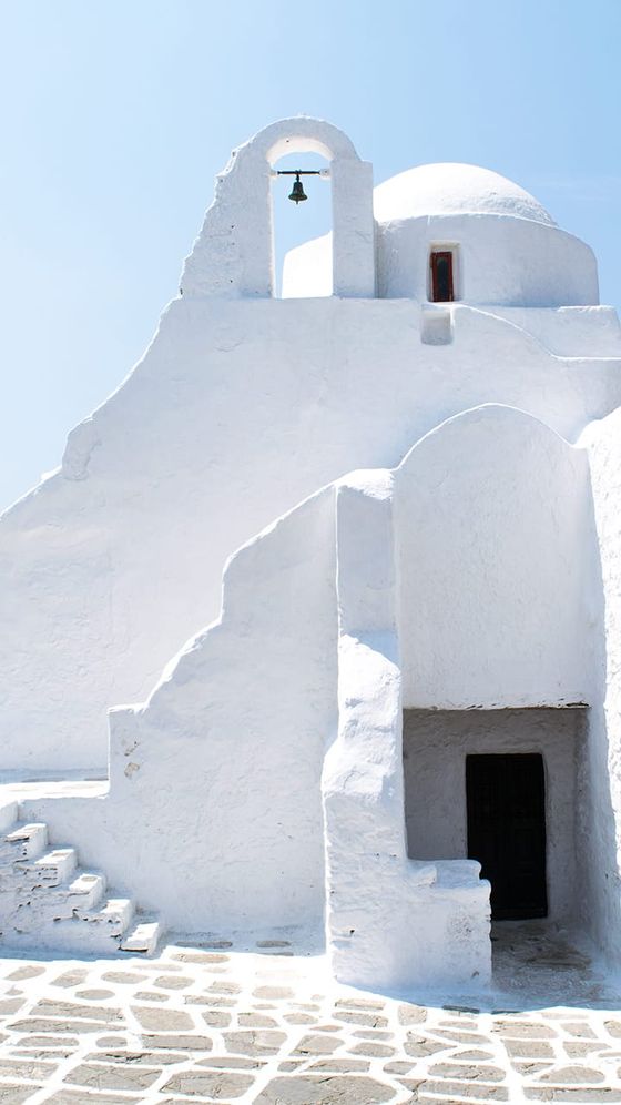 Whitewashed, domed building with bell, bright sunlight, stone steps, and patterned walkway, under a clear blue sky. Iconic Greek architecture.