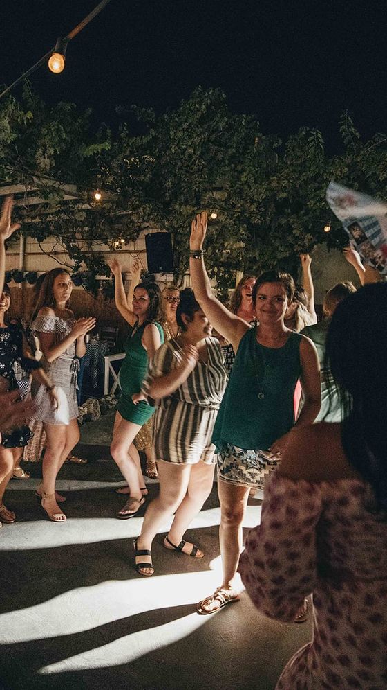 A group of people dancing joyfully under string lights at night, with trees in the background.