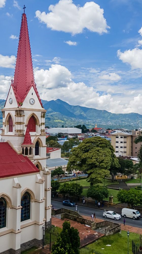 A red-roofed church with a tall steeple stands among trees and buildings, with lush mountains and a blue, cloudy sky in the background.