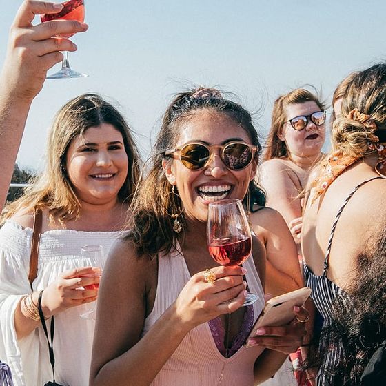 A group of people smiling and holding glasses of red wine at an outdoor gathering on a sunny day.