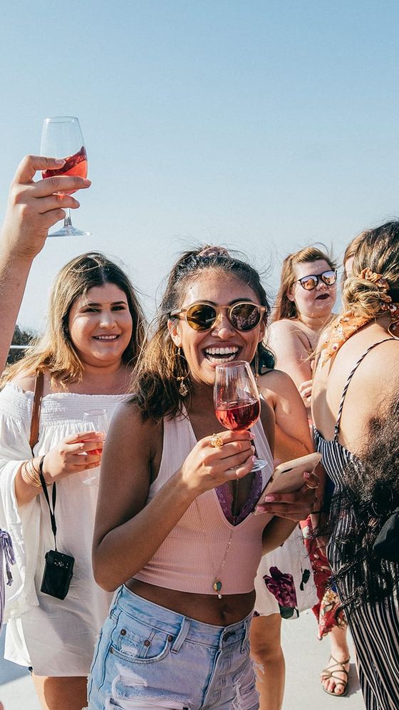 A group of people smiling and holding glasses of red wine at an outdoor gathering on a sunny day.