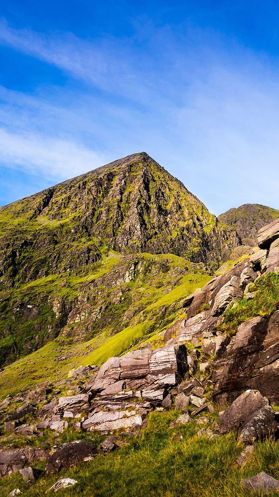 A rocky, green-covered mountain peak under a bright blue sky with scattered clouds, surrounded by grassy slopes and boulders.