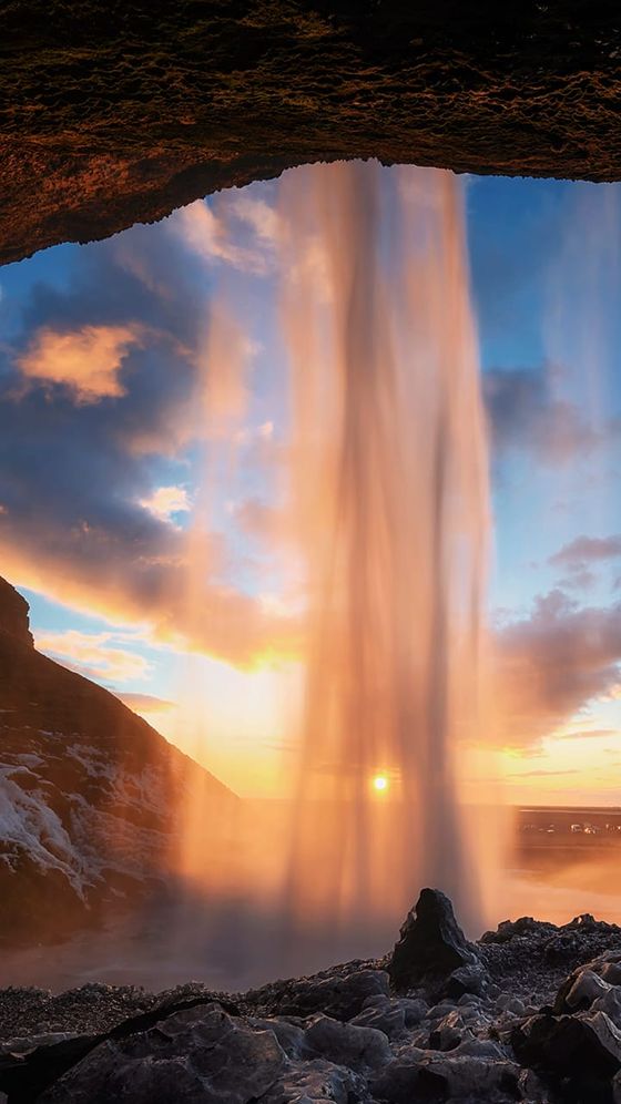 Waterfall cascading in front of a cave view, with a vibrant sunset and cloudy sky in the background.