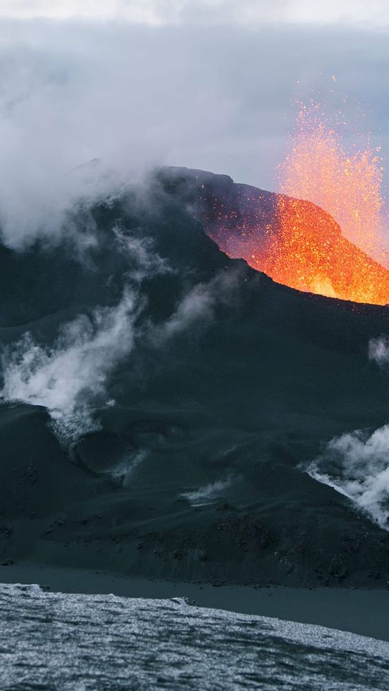 Active volcano erupting with glowing lava and ash, surrounded by smoke, under a cloudy sky.