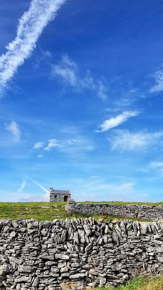 A small stone building stands on a grassy hill under a bright blue sky with scattered clouds and a contrail, bordered by a stone wall.