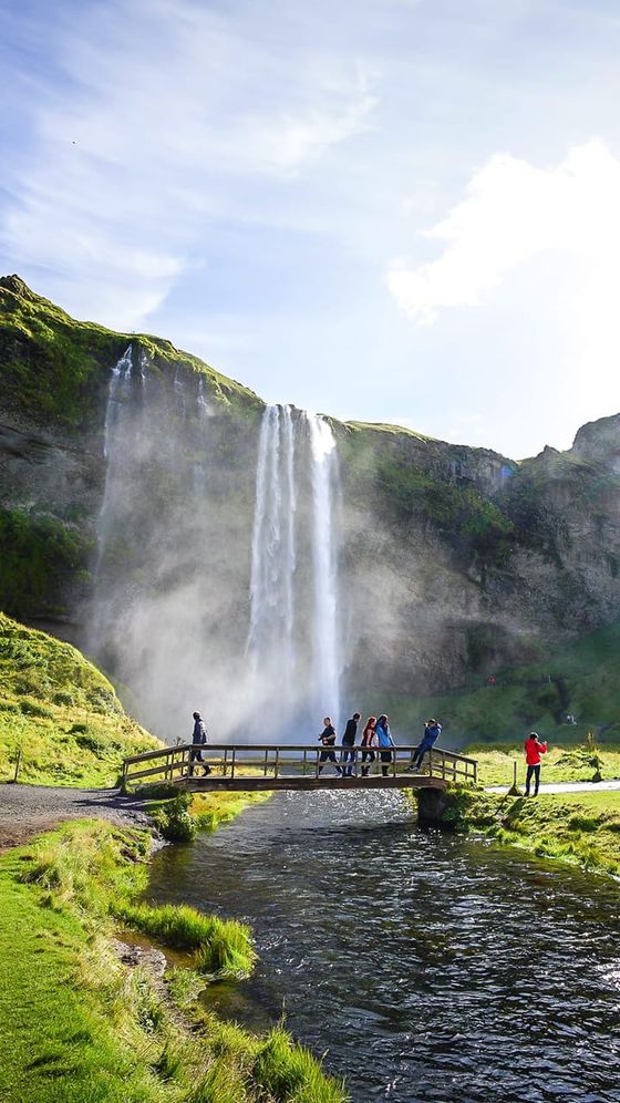 A group of people stand on a wooden bridge over a stream, with a tall waterfall and misty cliffs in the background under a blue sky.
