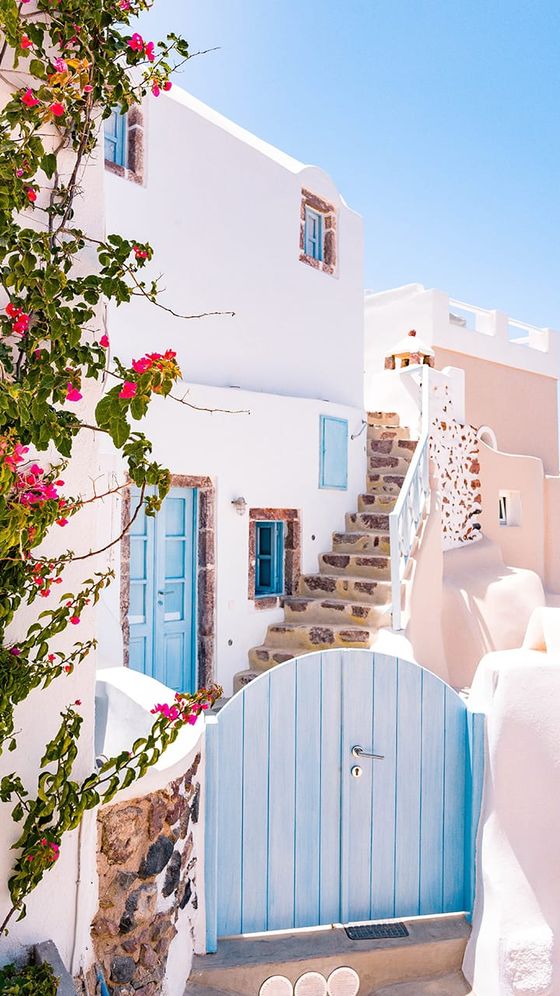 White Mediterranean house with pink flowers, blue doors, and a stone staircase under a bright blue sky.