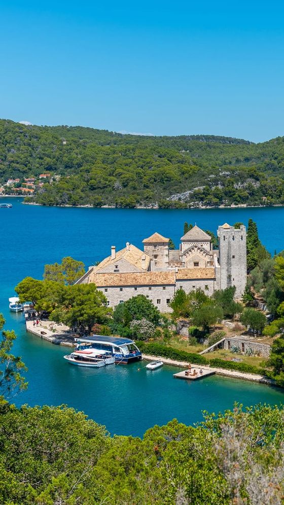 Scenic view of a historic stone monastery by a vibrant blue lake, surrounded by lush green hills, with boats docked nearby under a clear blue sky.