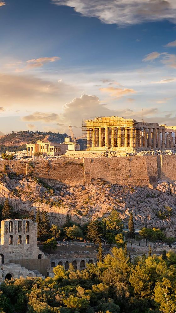 The Acropolis of Athens at sunset, with the Parthenon prominently atop a rocky hill, surrounded by lush greenery.