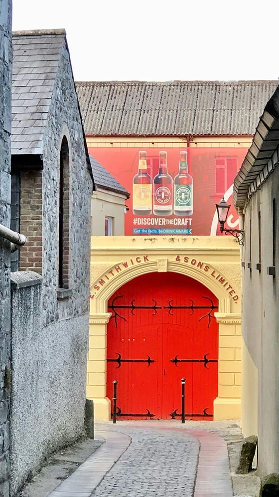 Narrow cobblestone alley leading to a vibrant red gate with "Smithwick & Sons Limited" above, flanked by stone and painted buildings.