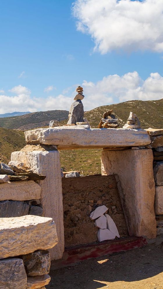 Ancient stone ruin with stacked rocks on top, set against a backdrop of rolling hills and a blue sky with fluffy clouds.