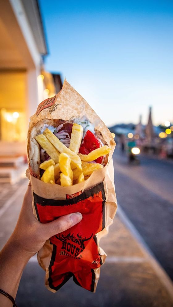 Person holding a paper-wrapped sandwich filled with fries, sauce, and toppings, with a blurred street and evening sky in the background.