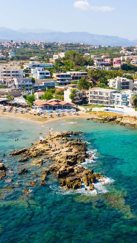 Aerial view of a coastal town with sandy beaches, rocky outcrops, turquoise water, and modern buildings surrounded by lush greenery.