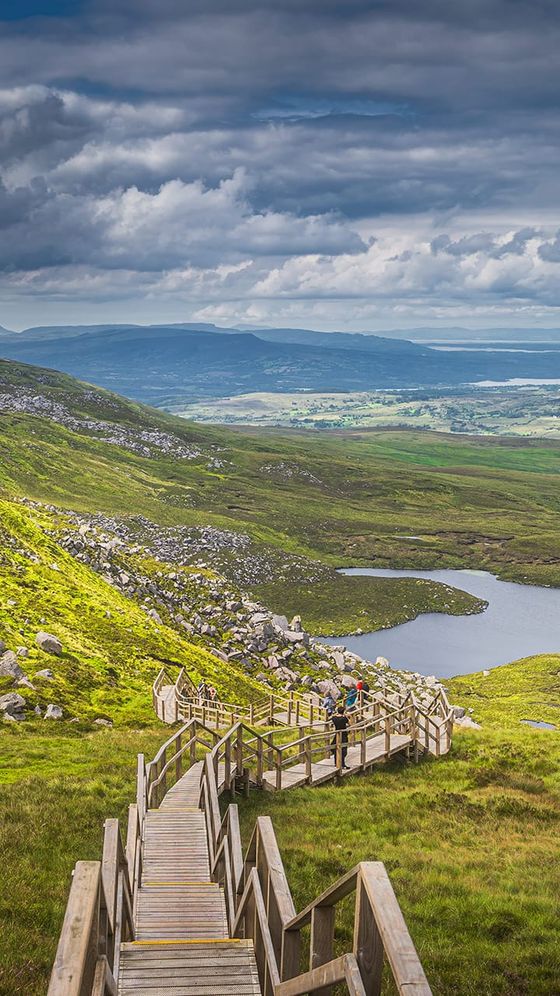 A wooden boardwalk leads down through green hills, overlooking a small lake and distant mountains under a partly cloudy sky.