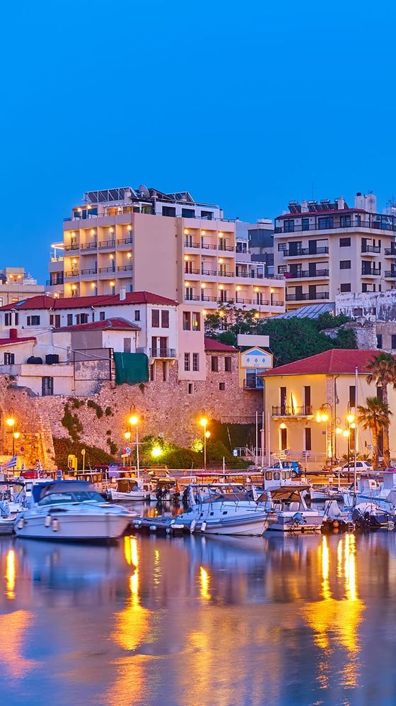 Scenic view of a marina at dusk with boats on calm water, buildings in the background, and warm lighting reflecting on the water.