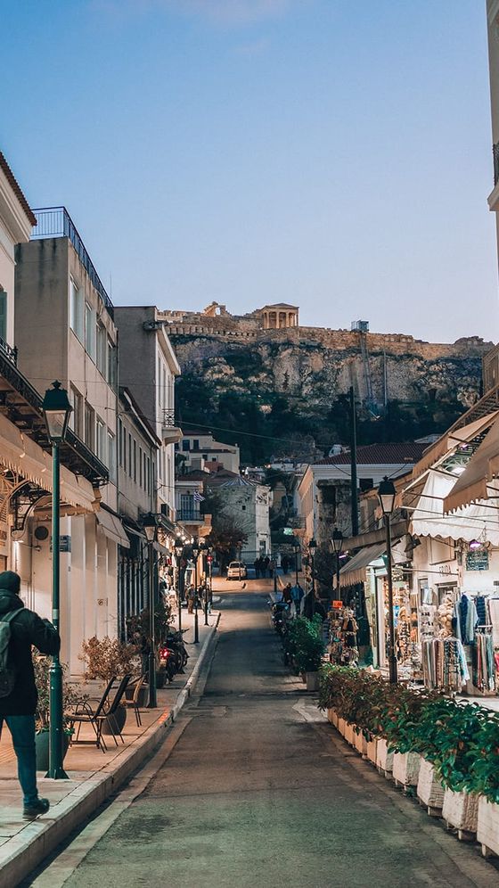 Narrow street lined with shops and potted plants, leading to the Acropolis under a twilight sky.