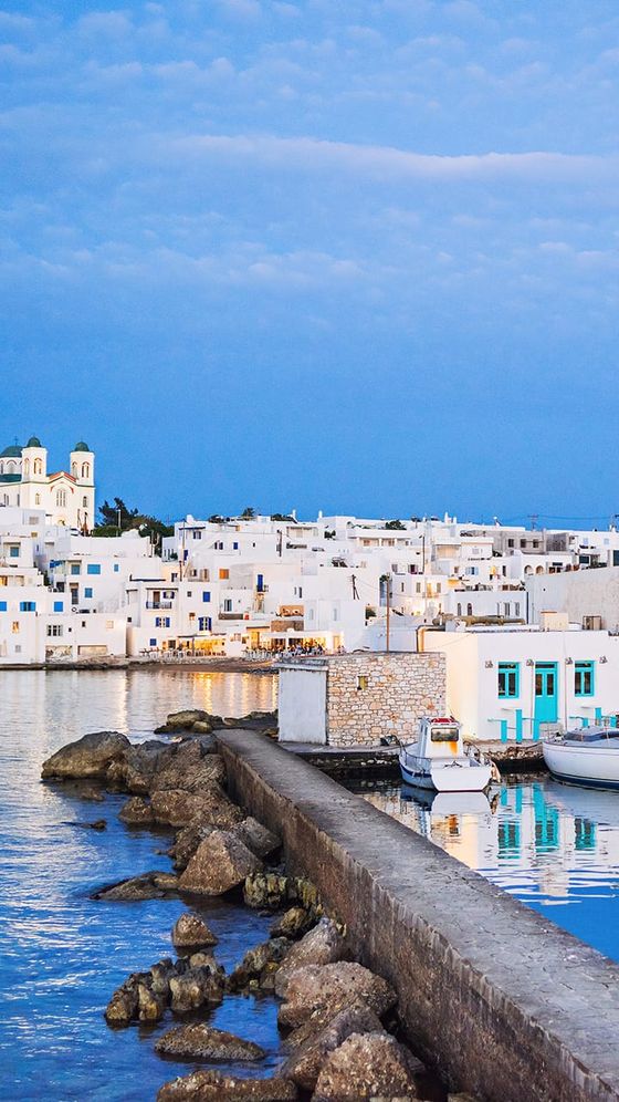 Coastal town with white buildings, small boats docked by the pier, and a blue sky at dusk, reflecting on calm waters.