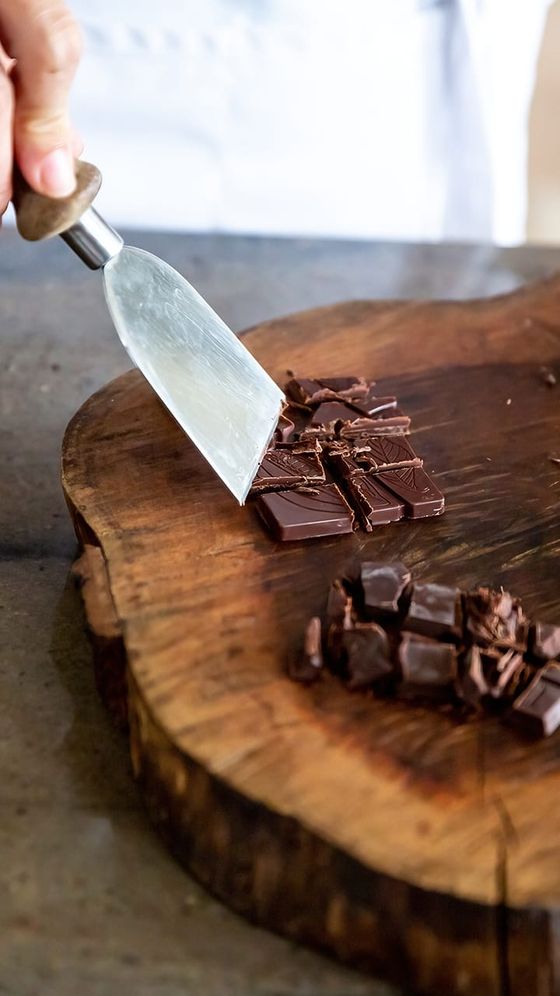 Hand using a scraper to chop dark chocolate on a round wooden board.