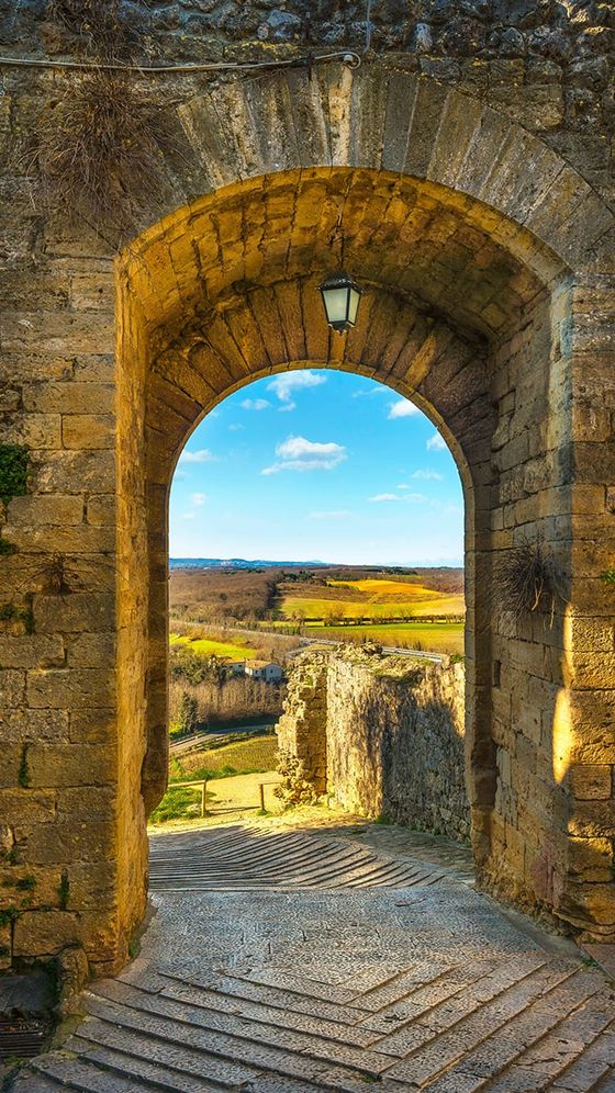 View of a vibrant landscape with fields and a clear blue sky through a stone archway, with a cobblestone path leading out.