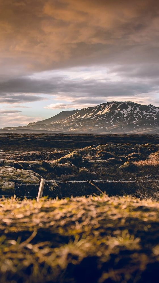 Scenic view of a snow-capped mountain under a dramatic sky, with a foreground of rocky, grassy terrain at sunset.