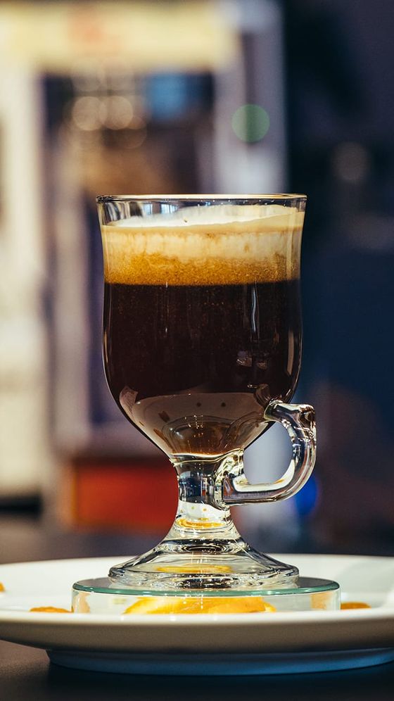 A glass mug of Irish coffee with a layer of frothy cream on top, set on a saucer with caramel drizzle, against a blurred background.