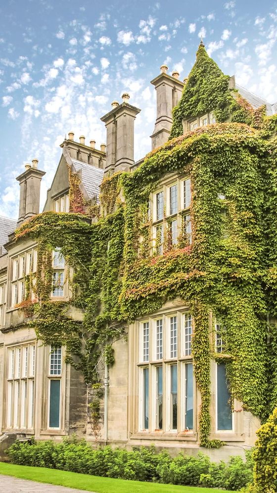 Historic stone building covered in green ivy, featuring tall chimneys and large windows, under a partly cloudy sky.