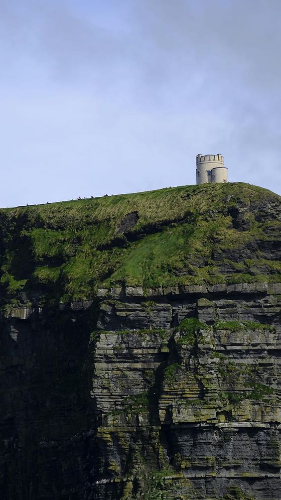 A small stone tower stands atop a grassy, cliffside hill, with rugged, vertical rock layers beneath and a cloudy sky above.