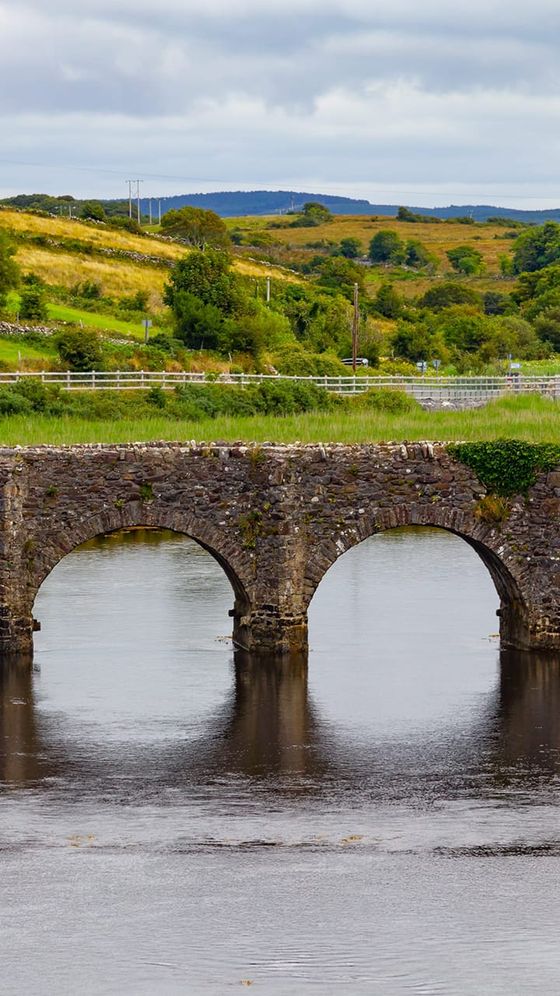 Stone bridge with two arches over a calm river, set in a lush green landscape with rolling hills and a cloudy sky.